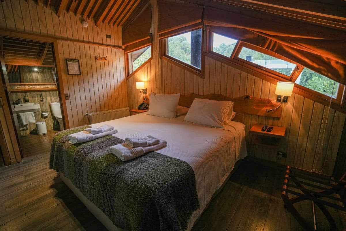 A wooden cabin bedroom with a bed, two bedside lamps, a wooden chair, and a window overlooking greenery.