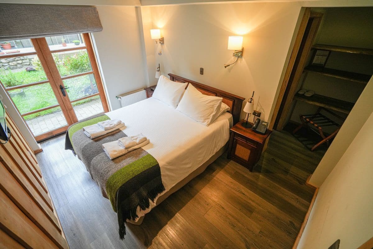 A bedroom with a large bed, wooden floor, two bedside lamps, a small table with a lamp, and a view of a garden through a sliding door.