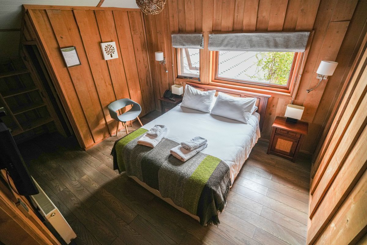 A wooden-paneled bedroom with a bed, a chair, a nightstand, and a window with a view of greenery.