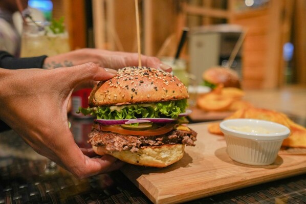 Una persona sosteniendo una hamburguesa con pan de sésamo, lechuga, tomate y pepinillos.