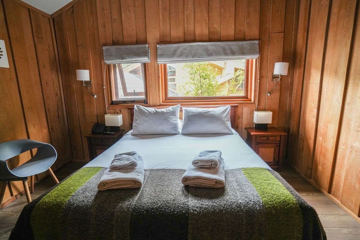A wooden-paneled room with a bed, two bedside lamps, a chair, and a window with a view.