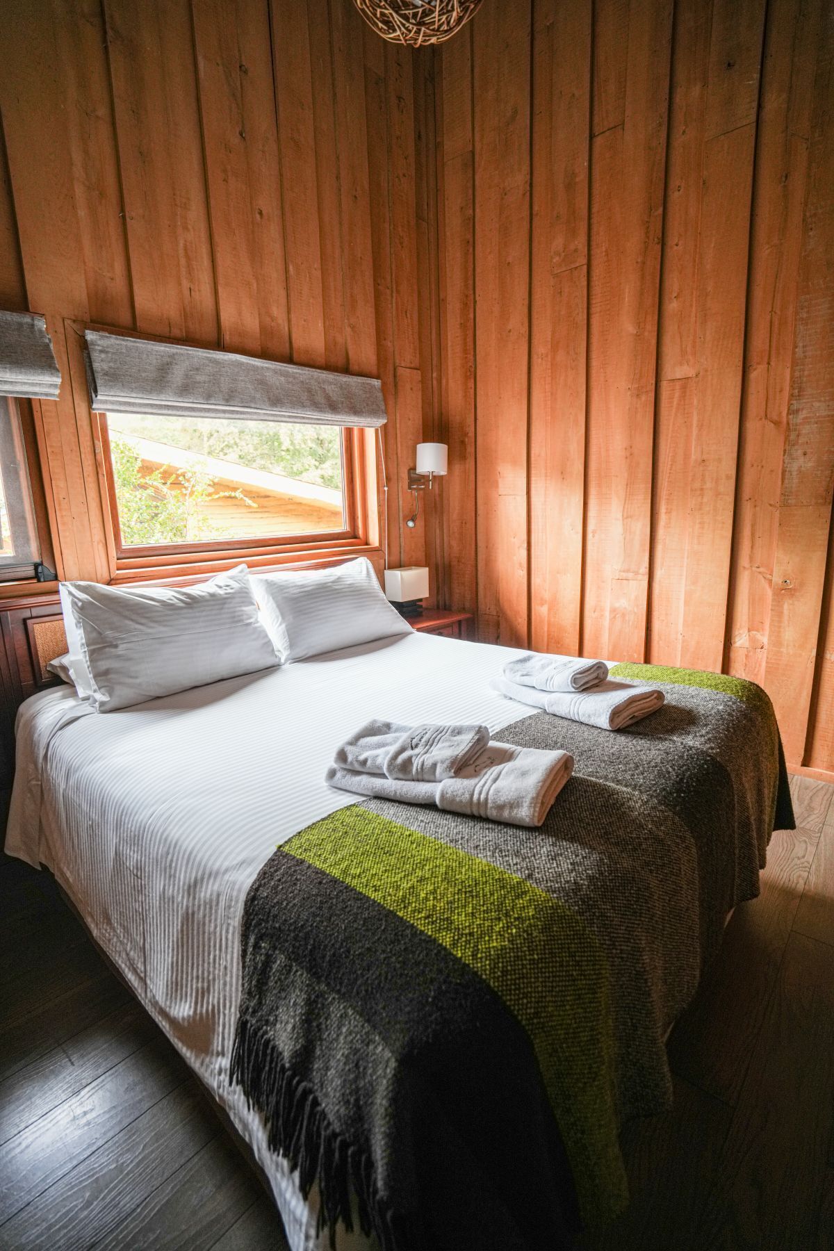 A wooden-paneled room with a bed, a window, and a wall-mounted lamp.