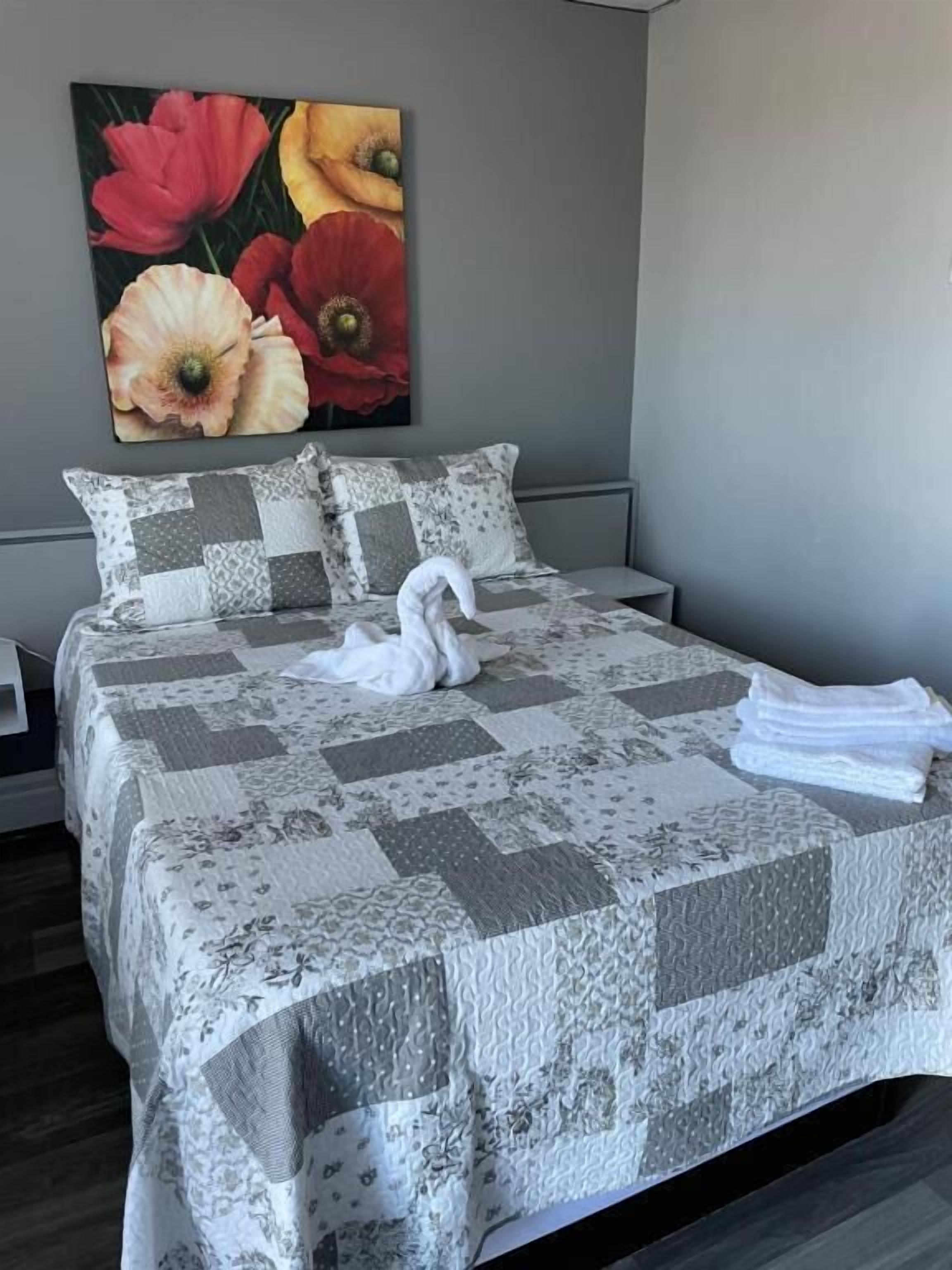 Een net opgemaakt bed met een geruit sprei en kussens, een witte zwaan decoratie en een bloemen schilderij aan de muur.