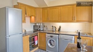 A compact kitchen with wooden cabinets, a built-in oven, a microwave, a washing machine, and a wine bottle on the counter. - Haven Cottage (Seahouses)