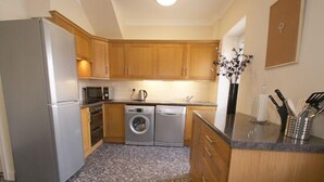 A compact kitchen with wooden cabinets, a stainless steel refrigerator, a washing machine, and a microwave. - Haven Cottage (Seahouses)