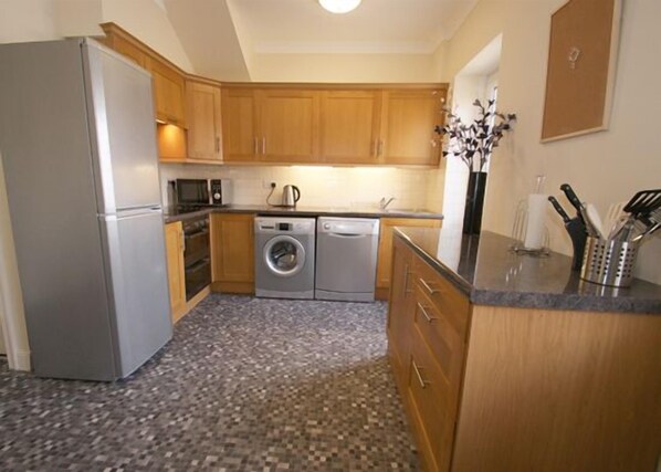 A compact kitchen with wooden cabinets, a stainless steel refrigerator, a washing machine, and a microwave.