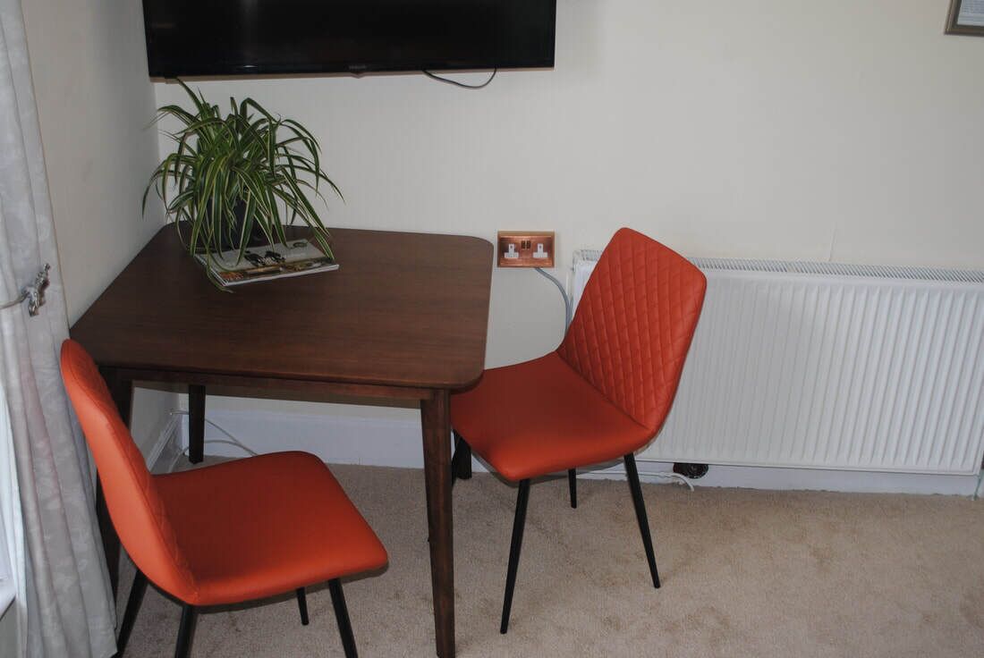 A small dining area with a wooden table, two orange chairs, a potted plant, and a television mounted on the wall.
