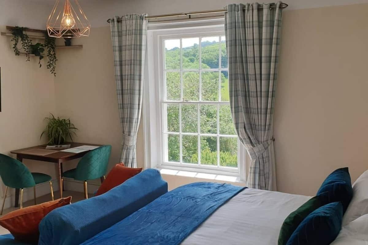 Ein Schlafzimmer mit einem großen Bett, zwei Nachttischen, einem Schreibtisch und einem Stuhl. Es gibt ein Fenster mit Vorhängen und eine Pendelleuchte.