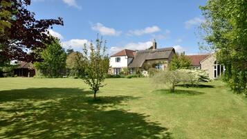 A residential property with a large lawn, a thatched-roof house, and a white building with a red roof.
