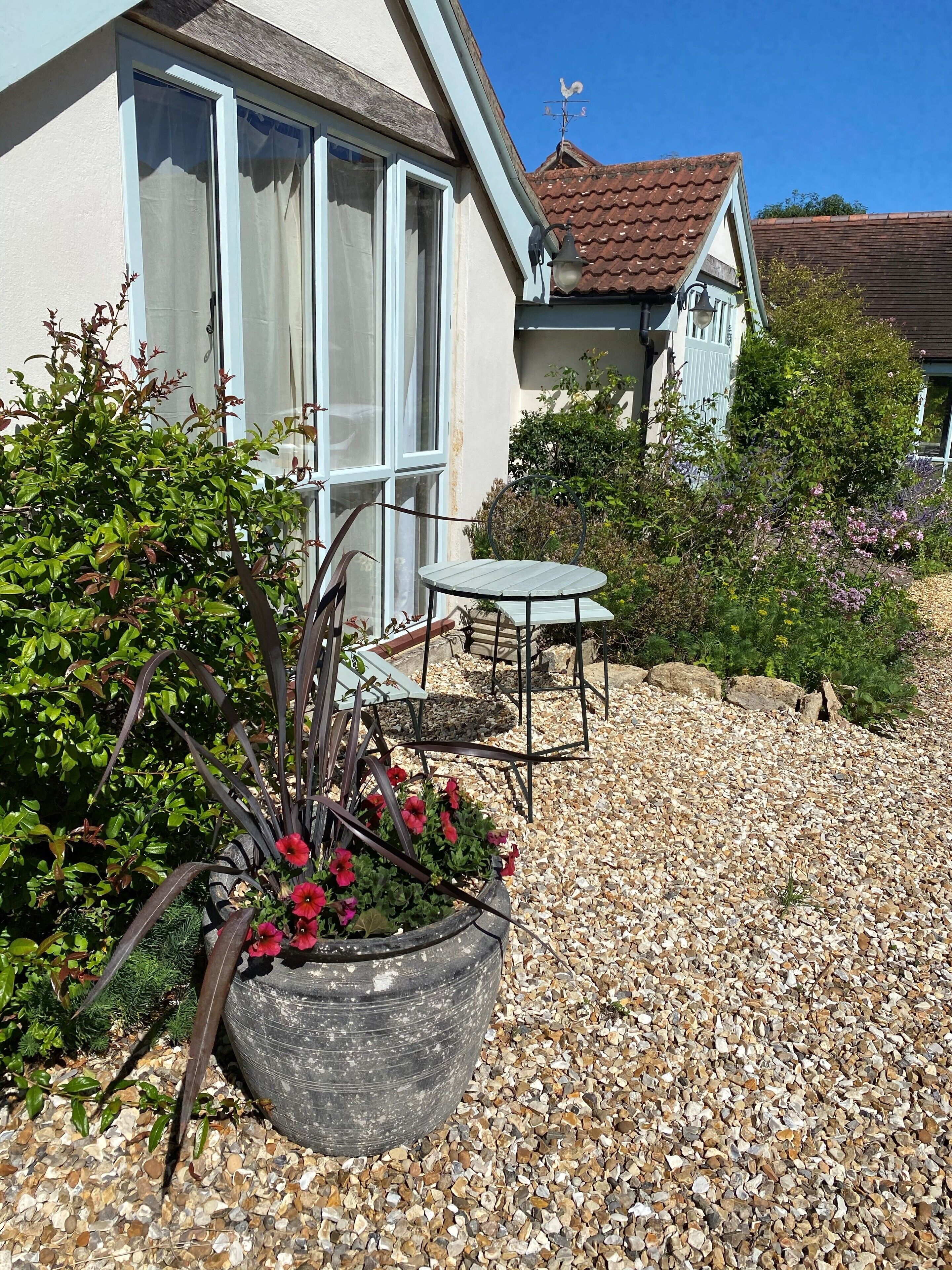 A house with a gravel patio, a metal table and chair, and a large planter with red flowers.