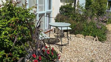A house with a gravel patio, a metal table and chair, and a large planter with red flowers.