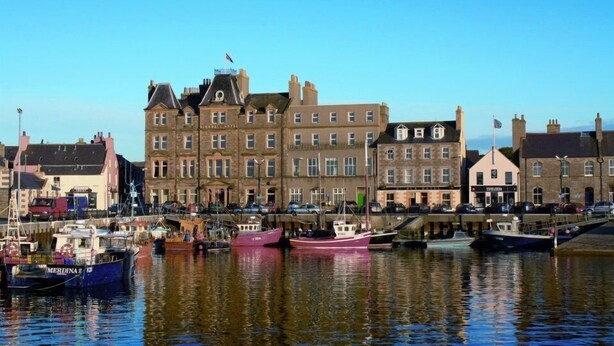 A waterfront hotel with multiple stories, a flag on top, and boats docked in the water. - St Ola Hotel (Kirkwall)