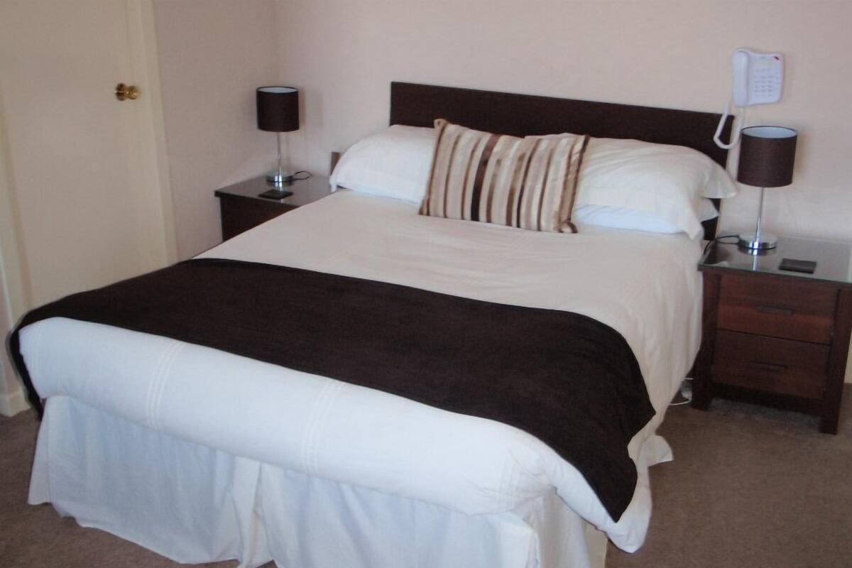 A neatly made bed with a white and brown bedspread, two bedside lamps, a bedside phone, and a small nightstand.