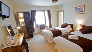 A hotel room with two beds, a desk with a computer and a tray of items, a TV, and a window with curtains. - St Ola Hotel (Kirkwall)