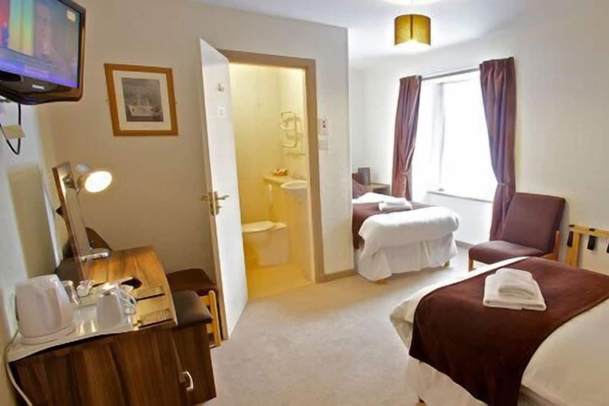 A hotel room with a bed, a chair, a desk with a lamp, a TV, and a bathroom visible through an open door.
