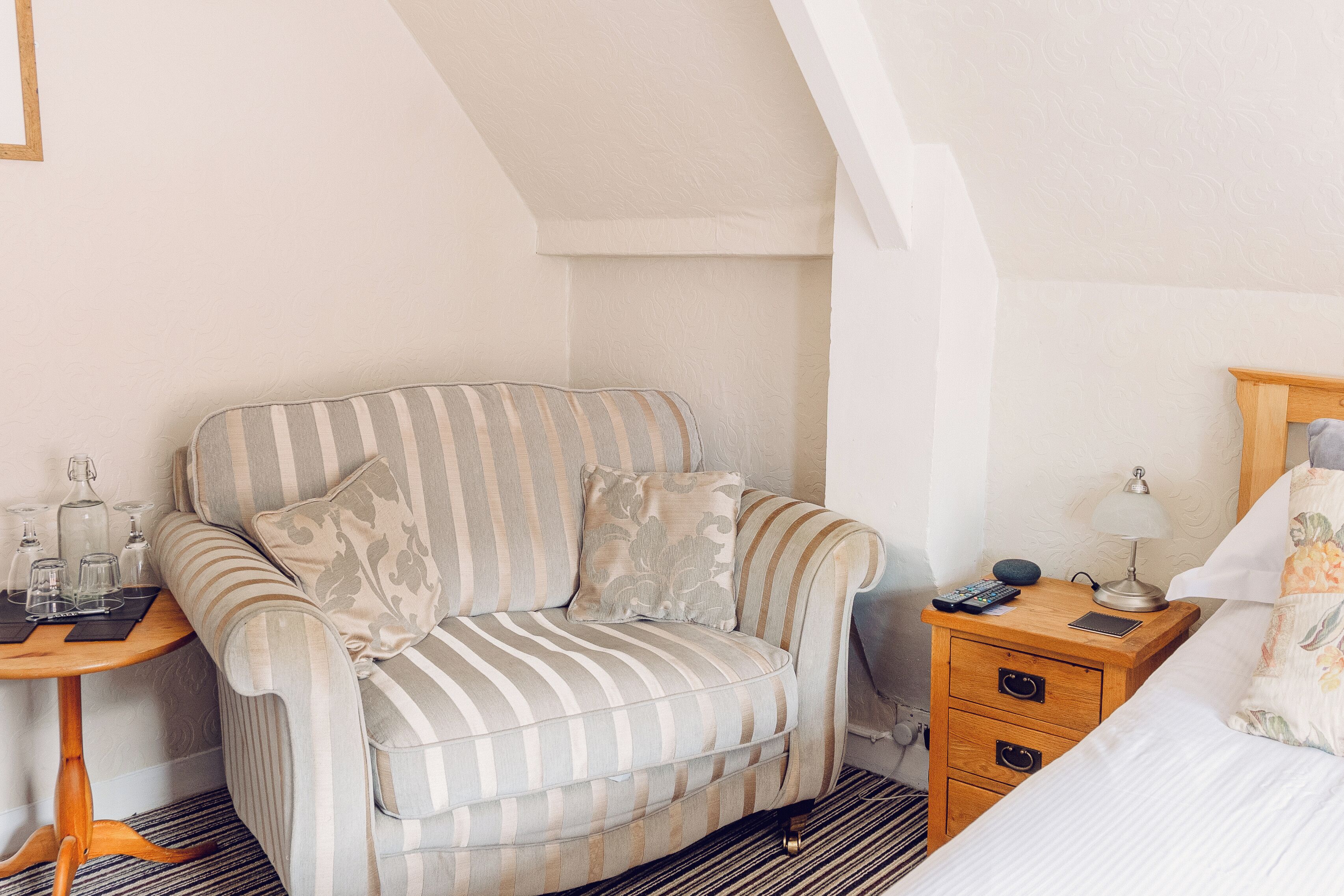 Een gezellige kamer met een gestreept bankje, een klein houten bijzettafeltje met een lamp, en een bed met bloemenbeddengoed.