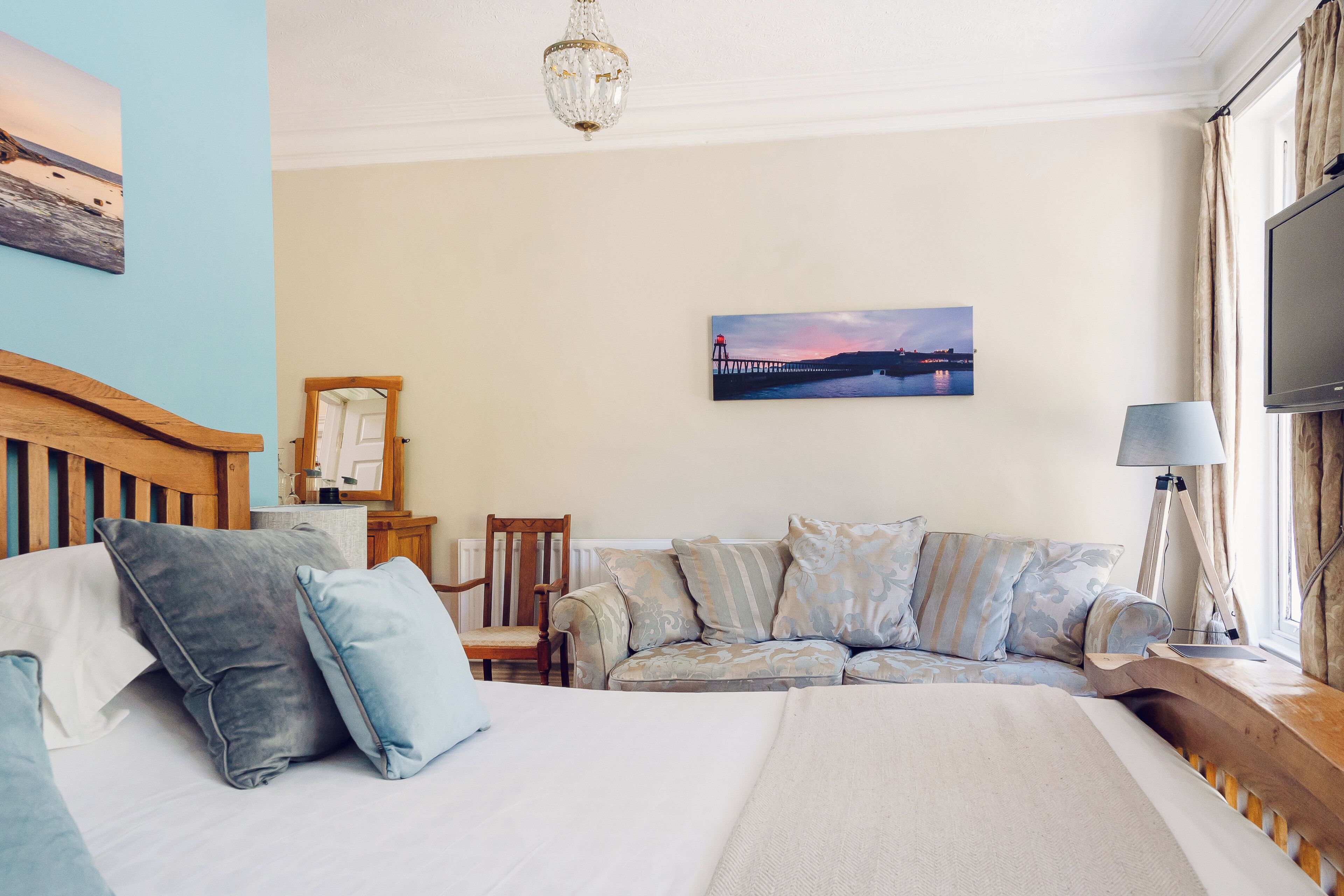 Ein Schlafzimmer mit Bett, Holz-Kopfteil, Sofa, Sessel, Schreibtisch, Lampe, Fernseher und einem Bild an der Wand.