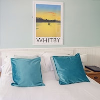 Et hotelværelse med en seng, to puder, et natbord, en lampe og et billede i ramme af en solnedgang over vandet, med ordet "WHITBY" under.