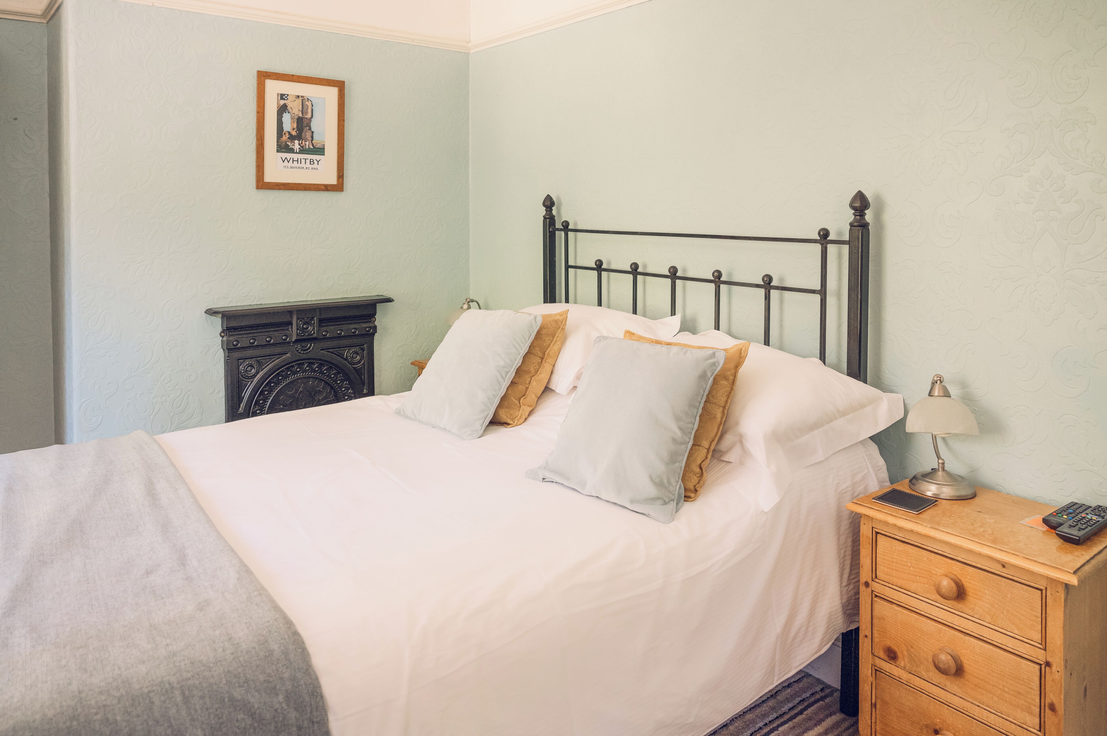 A neatly made bed with white linens and two pillows, a bedside table with a lamp and a remote control, and a framed picture on the wall.