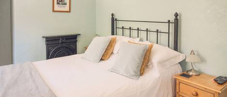 A neatly made bed with white linens and two pillows, a bedside table with a lamp and a remote control, and a framed picture on the wall.
