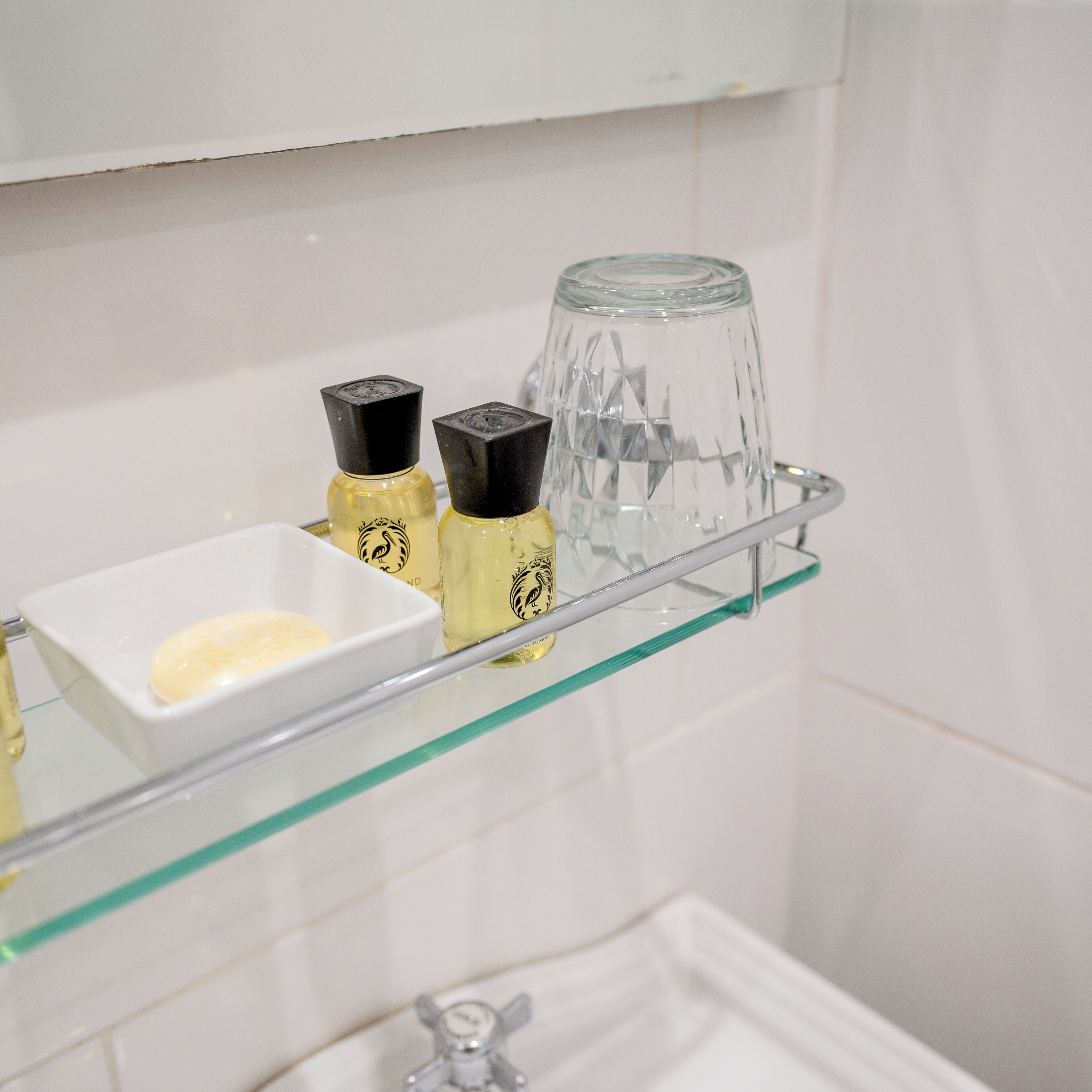A bathroom shelf with a glass container, two bottles with black caps, and a small white dish with a bar of soap.