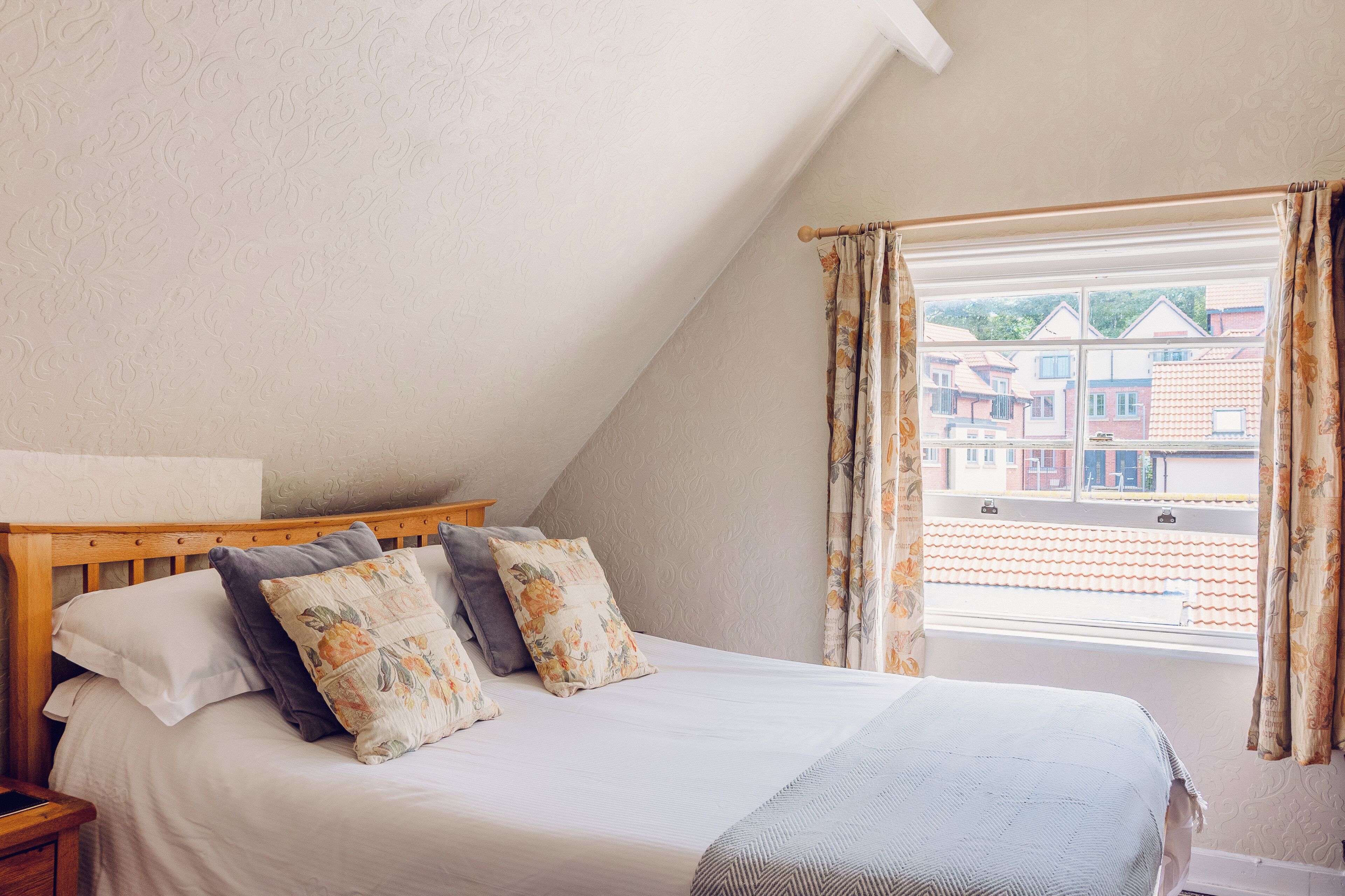 A bedroom with a sloped ceiling, a wooden headboard, a bed with white and floral pillows, and a window with curtains.