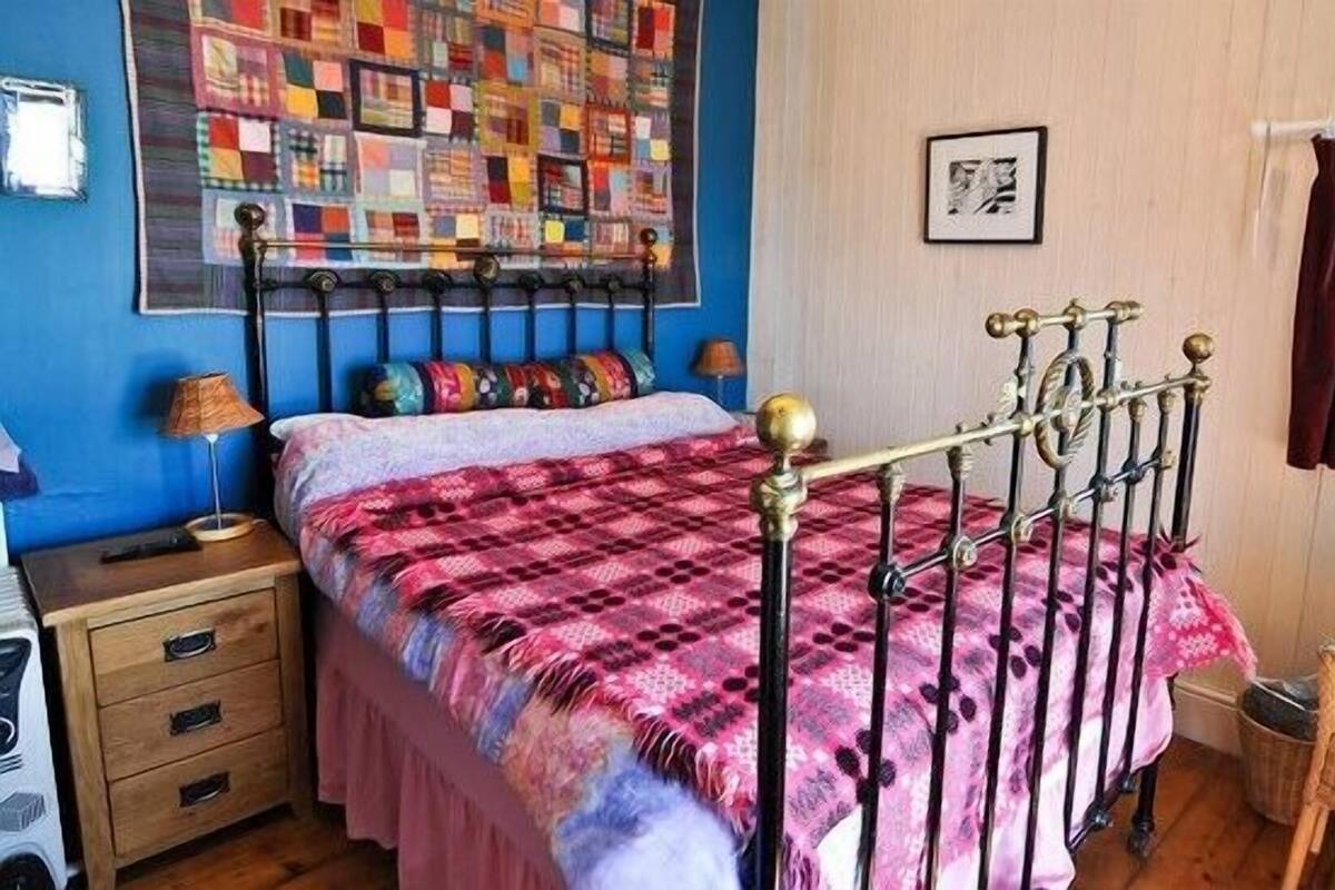 A bedroom with a bed, a nightstand, a chair, a quilt, and a wall with a picture.
