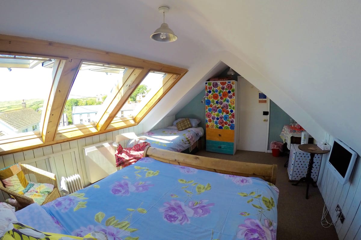 A bedroom with a sloped ceiling, a bed with floral bedding, a wardrobe with a colorful floral pattern, and a small table with a polka dot tablecloth.