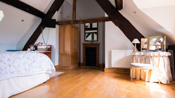 A bedroom with a wooden floor, a bed with a floral bedspread, a wooden wardrobe, a fireplace, a radiator, and a vanity with a mirror. - Huntlands Farm (Worcester)