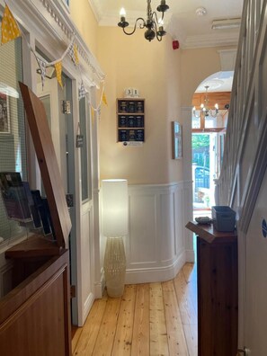 A hallway with wooden flooring, a chandelier, a wall-mounted shelf with jars, and a door with a calendar. - The Courtyard Guest House (Great Yarmouth)