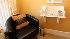 A room with a black leather sofa, a small trash bin, a white shelf with a coffee machine, cups, and a bowl of snacks.