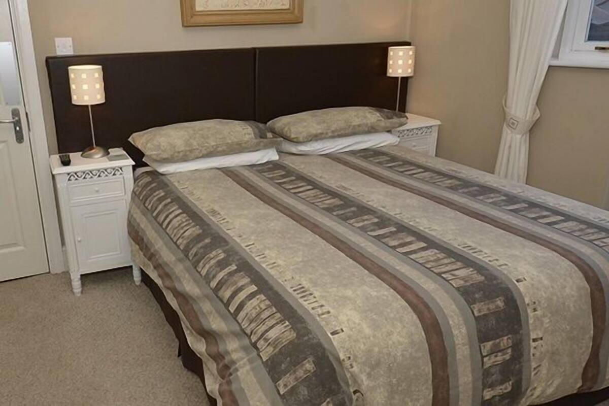 Een slaapkamer met een groot bed, twee nachtkastjes met lampen, een raam met gordijnen en een deur met een handgreep.