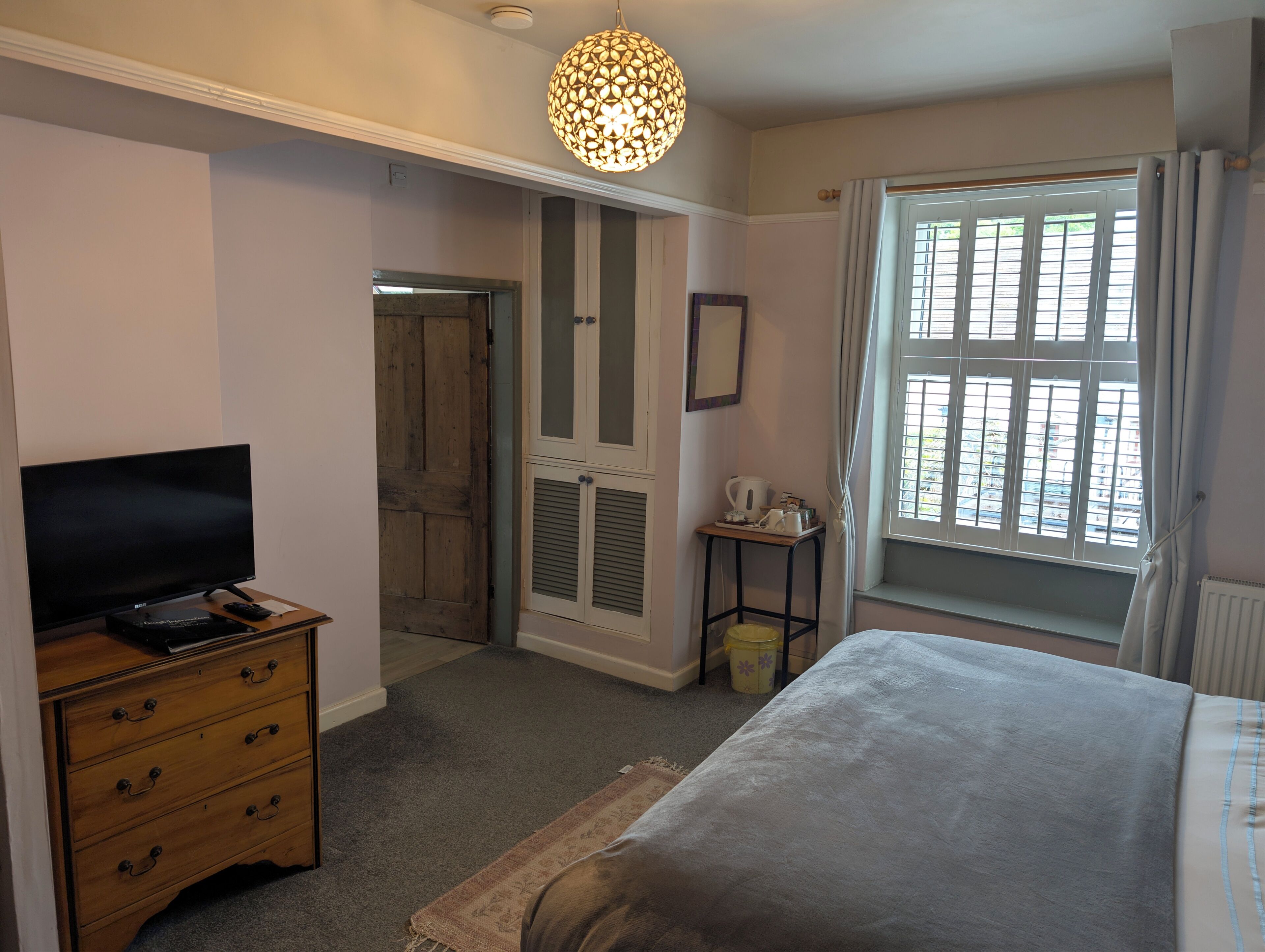 Un dormitorio con cama, cómoda, televisor, ventana con cortinas y puerta que da a otra habitación.