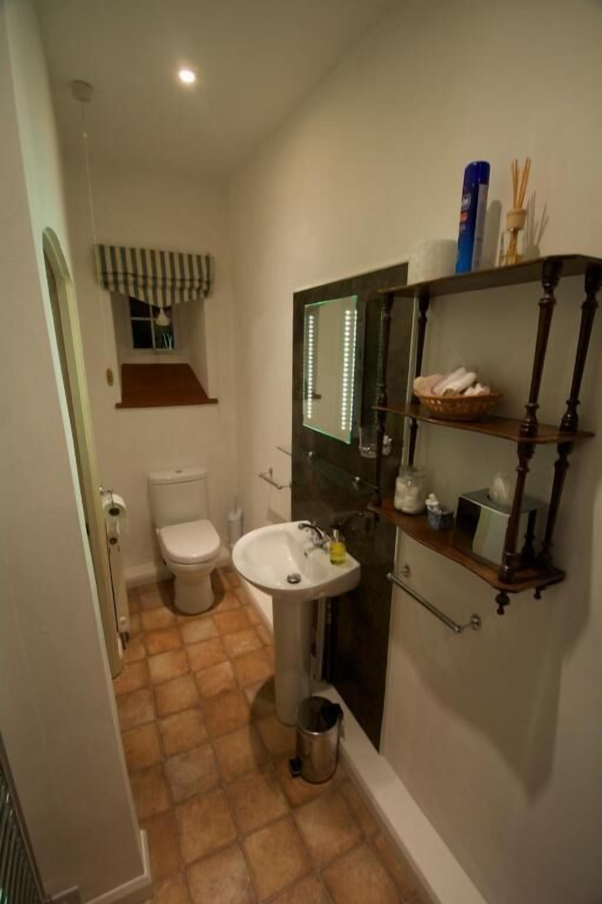 A bathroom with a toilet, sink, and mirror. There is a shelf with toiletries and a trash can.