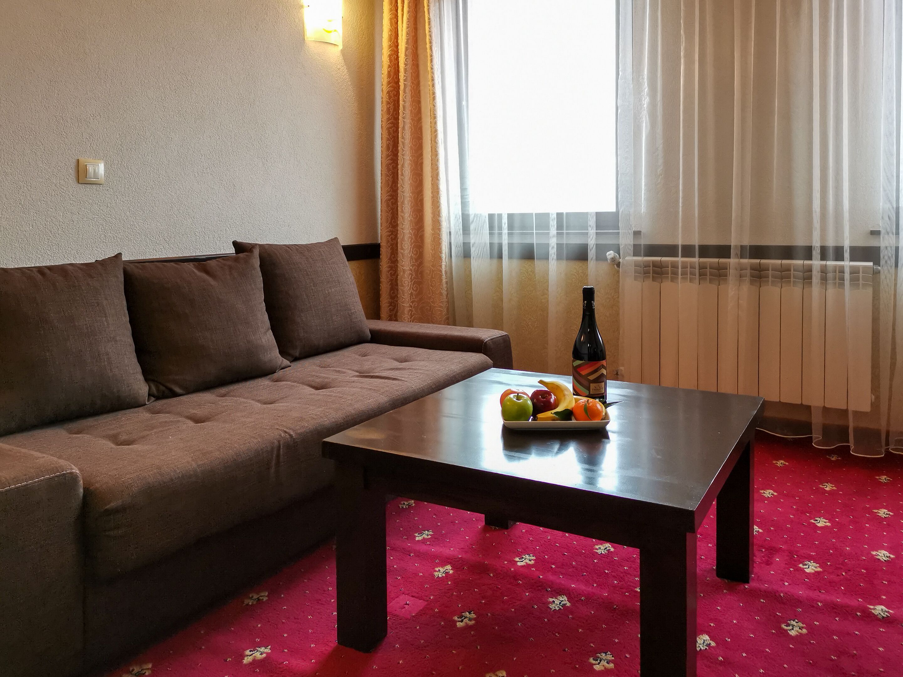 A living room with a brown sofa, a wooden coffee table with fruits and a bottle, and a red carpet.