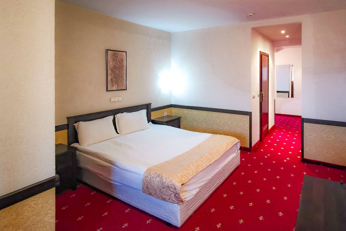 A hotel room with a bed, nightstands, a wall-mounted light, and a red carpet.