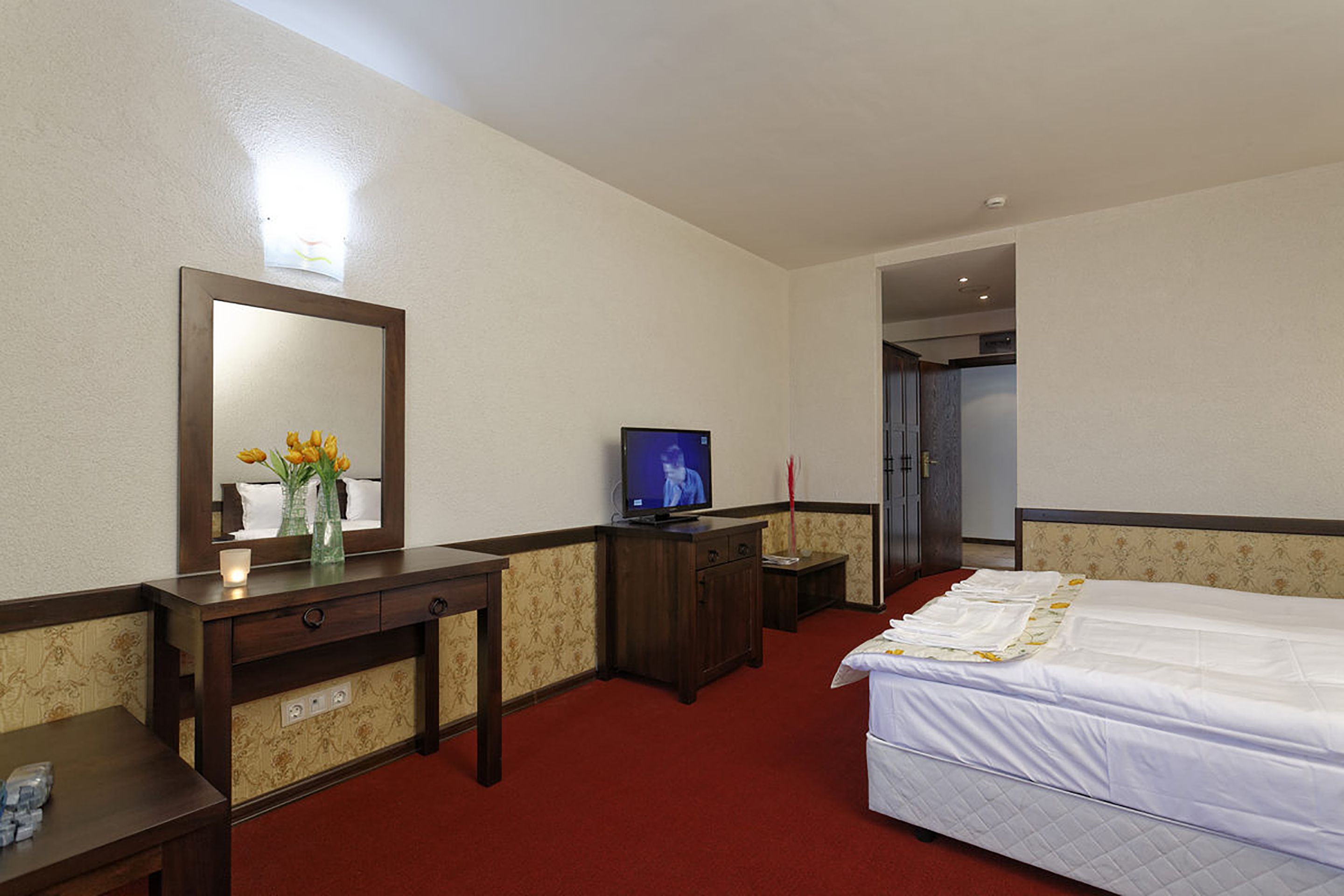 Ein Hotelzimmer mit einem Bett, einem Schreibtisch mit Fernseher, einem Spiegel und einer Vase mit Blumen.
