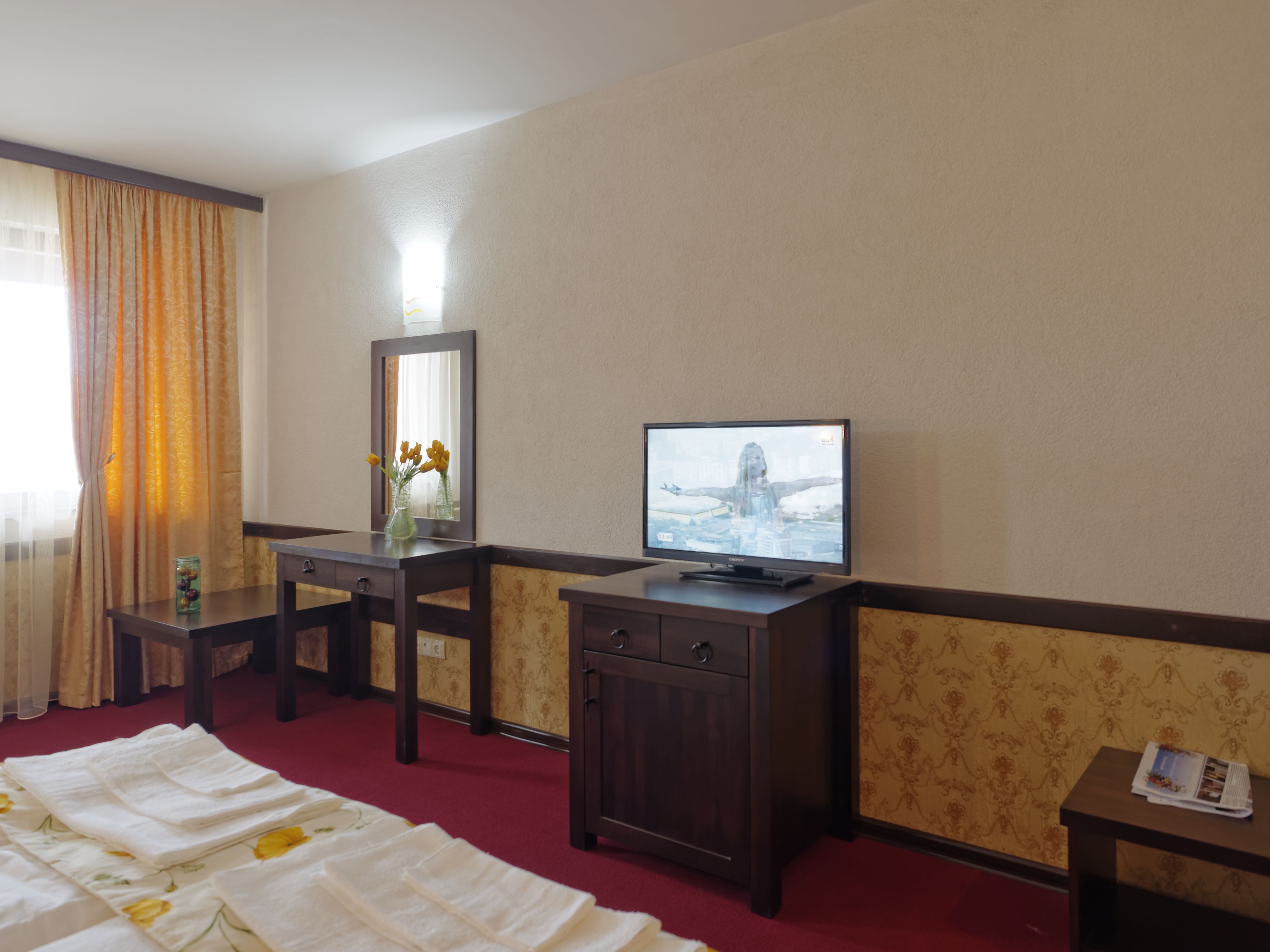 A hotel room with a bed, a desk with a television, a mirror, and a small table with a vase of flowers.
