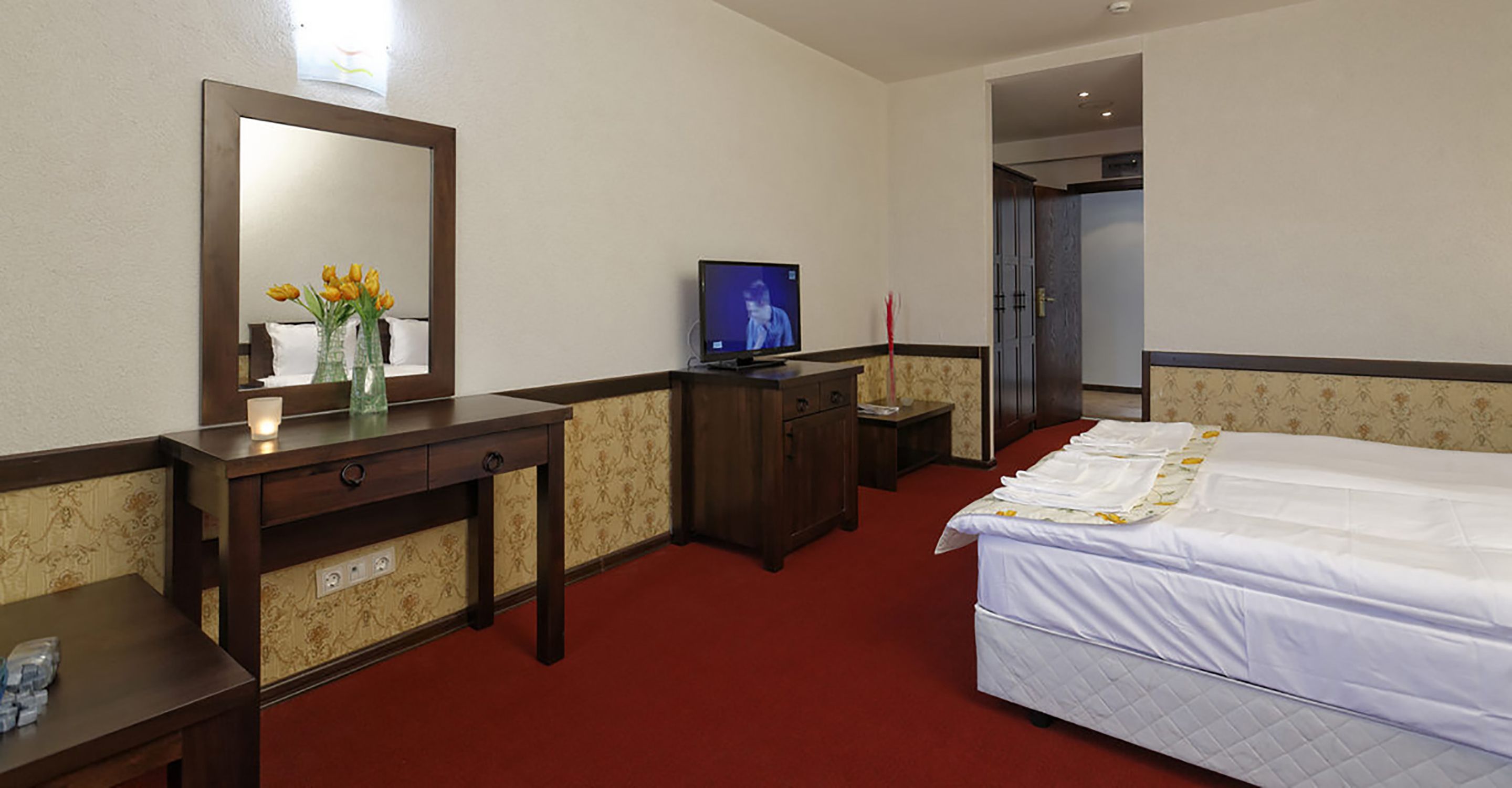 A hotel room with a bed, a desk, a television, a mirror, and a vase with flowers.