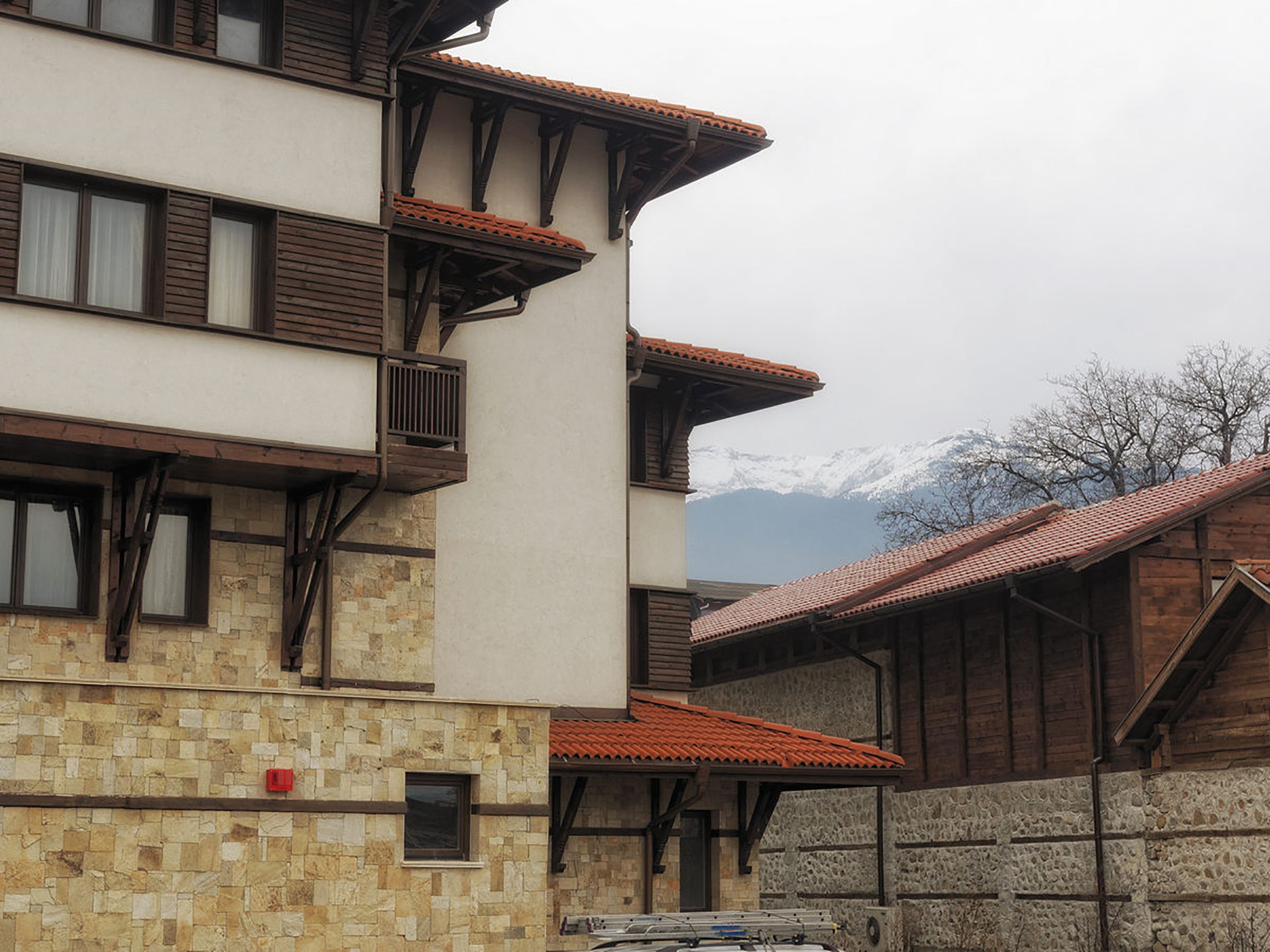 Un edificio de varios pisos con paredes exteriores de piedra y madera, balcones con techos de tejas rojas y un paisaje montañoso de fondo.