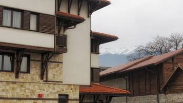 Un edificio de varios pisos con paredes exteriores de piedra y madera, balcones con techos de tejas rojas y un paisaje montañoso de fondo.