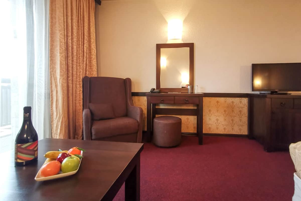 A hotel room with a brown leather chair, a small table with a fruit plate, a wooden desk with a mirror, a television, and a red carpet.