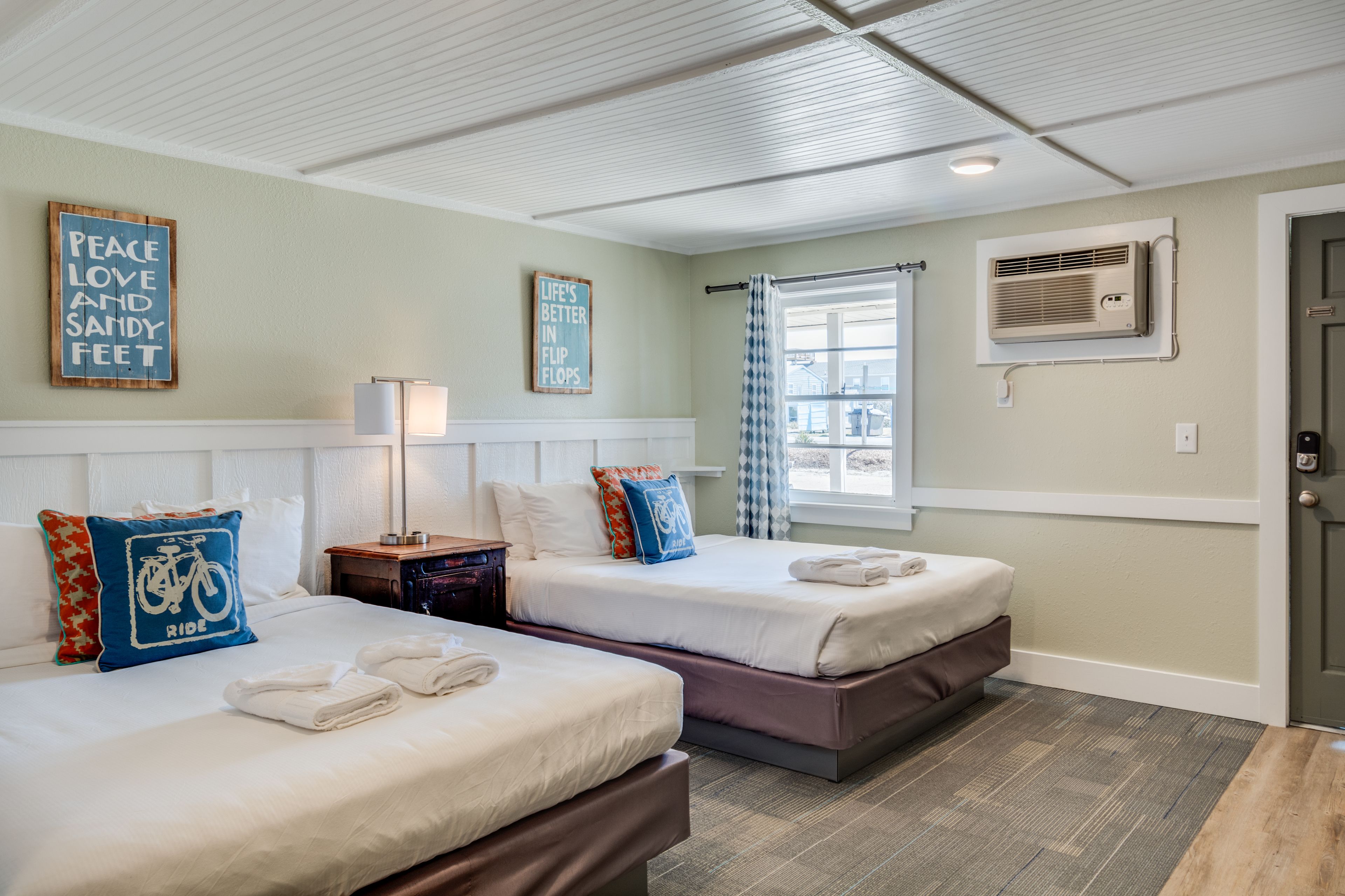 A hotel room with two beds, a window with a view, a wall-mounted air conditioner, and a door.