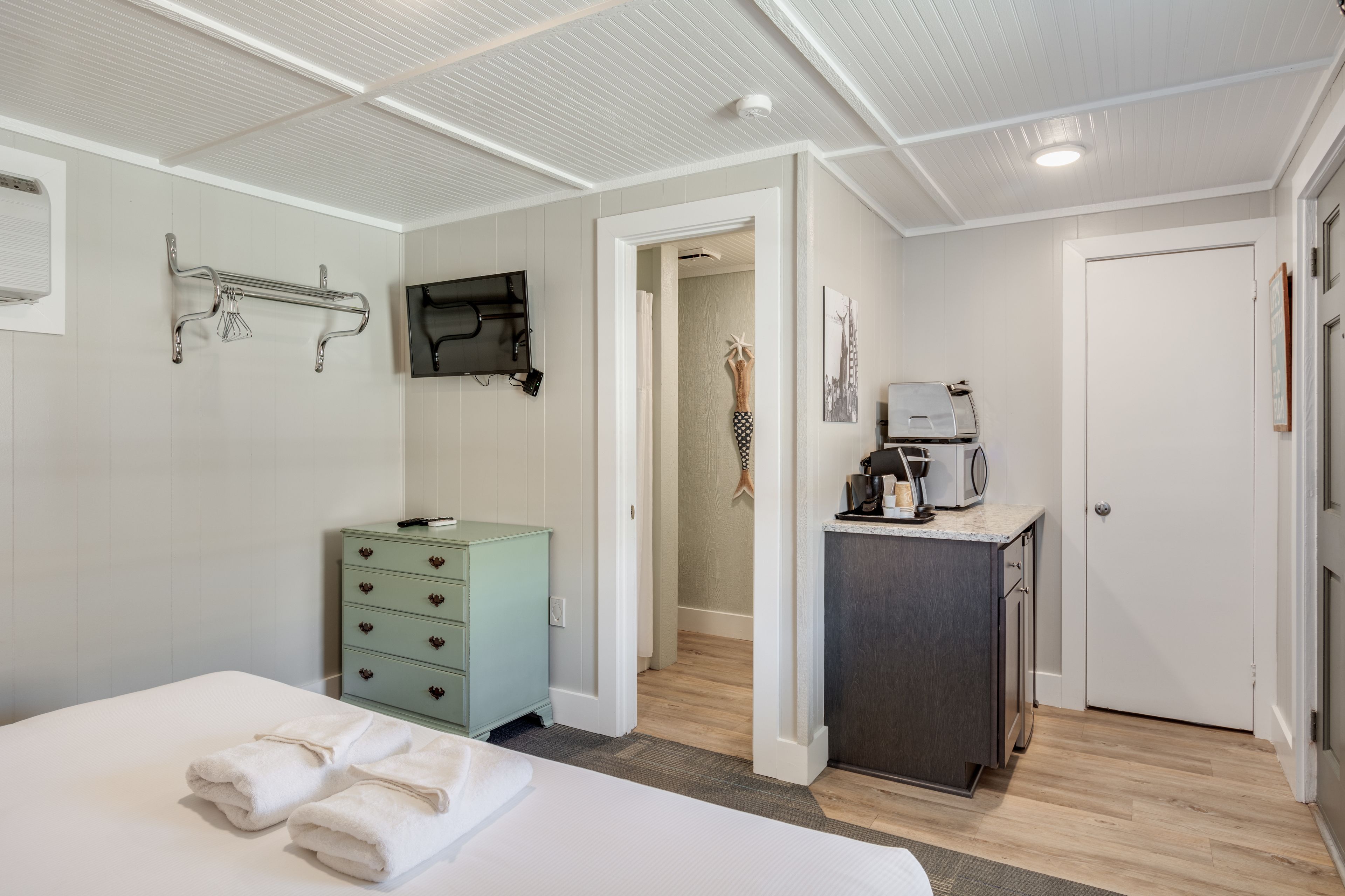 A hotel room with a bed, a green dresser, a desk with a coffee maker, and a TV mounted on the wall.