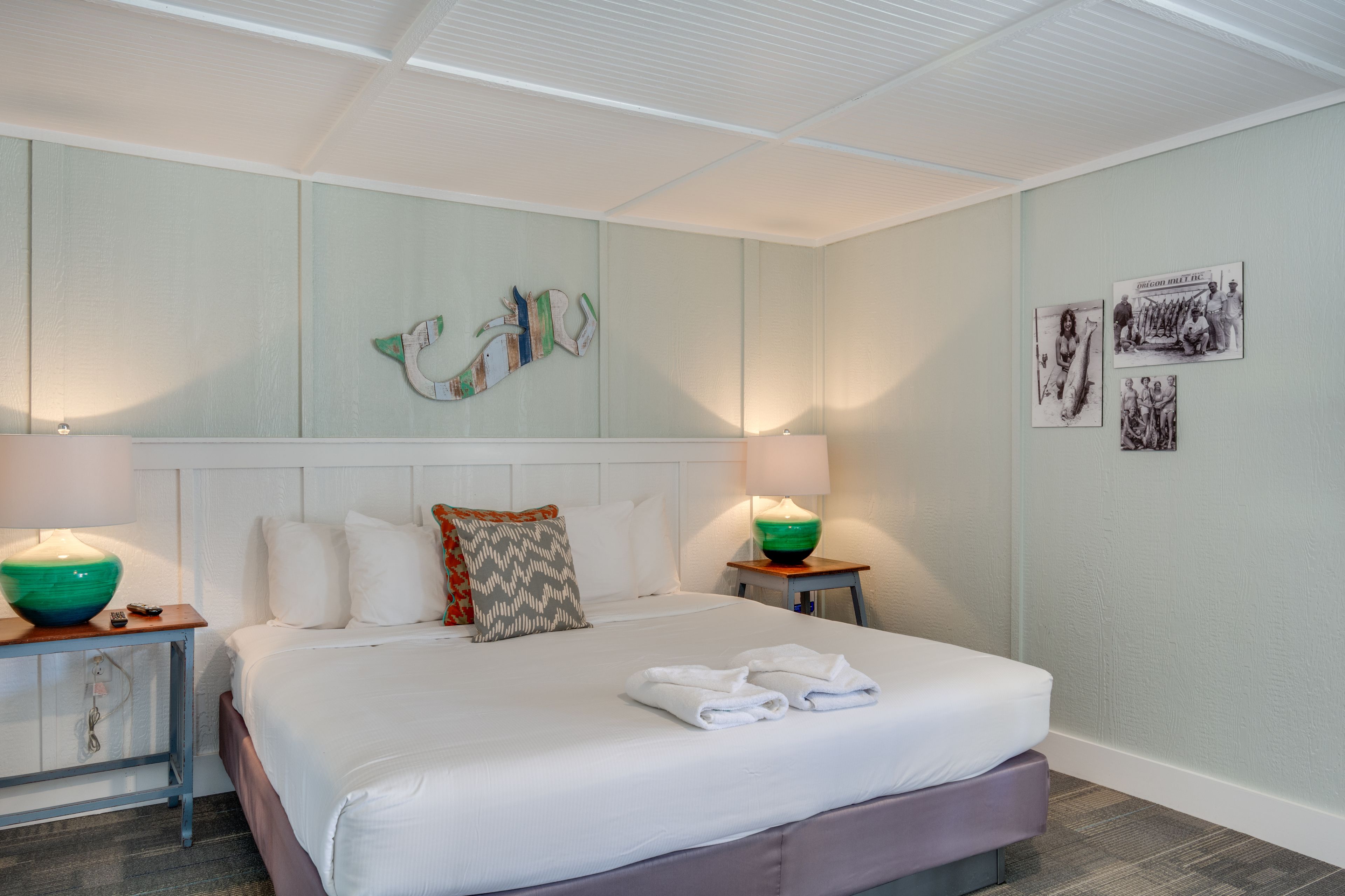 A neatly made bed with white linens and two pillows, a bedside table with a lamp, and a wall with framed pictures.