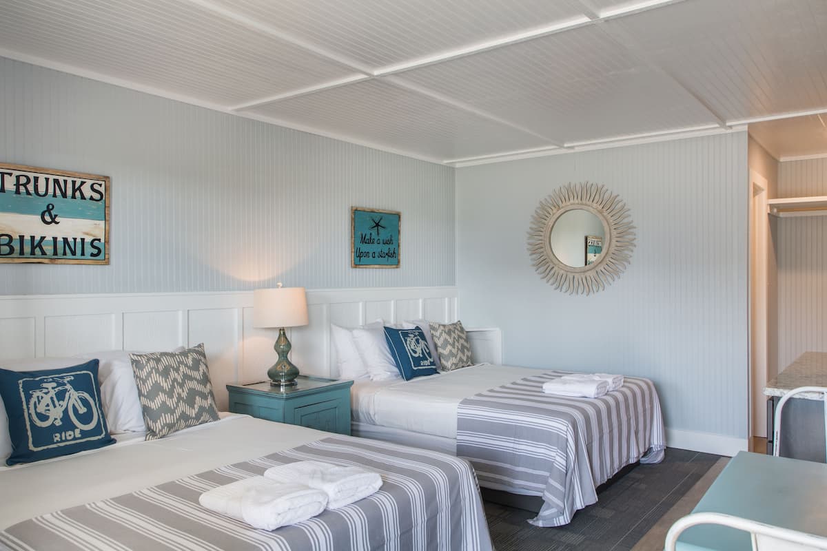 A hotel room with two beds, a mirror, a sign reading "Trunks & Bikinis", and a wall clock.