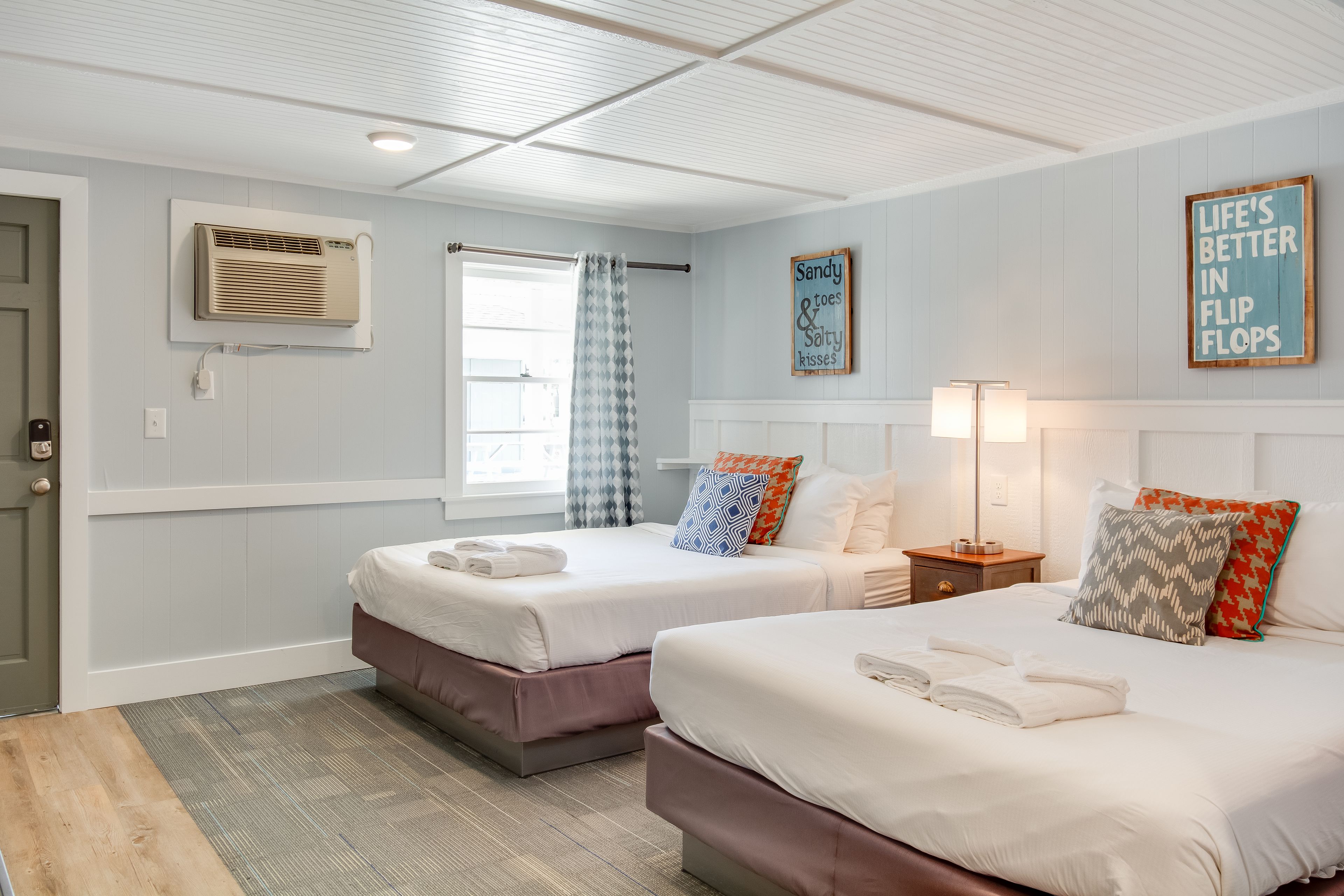 A hotel room with two beds, a window with a curtain, an air conditioning unit, a bedside table with a lamp, and a wall sign.