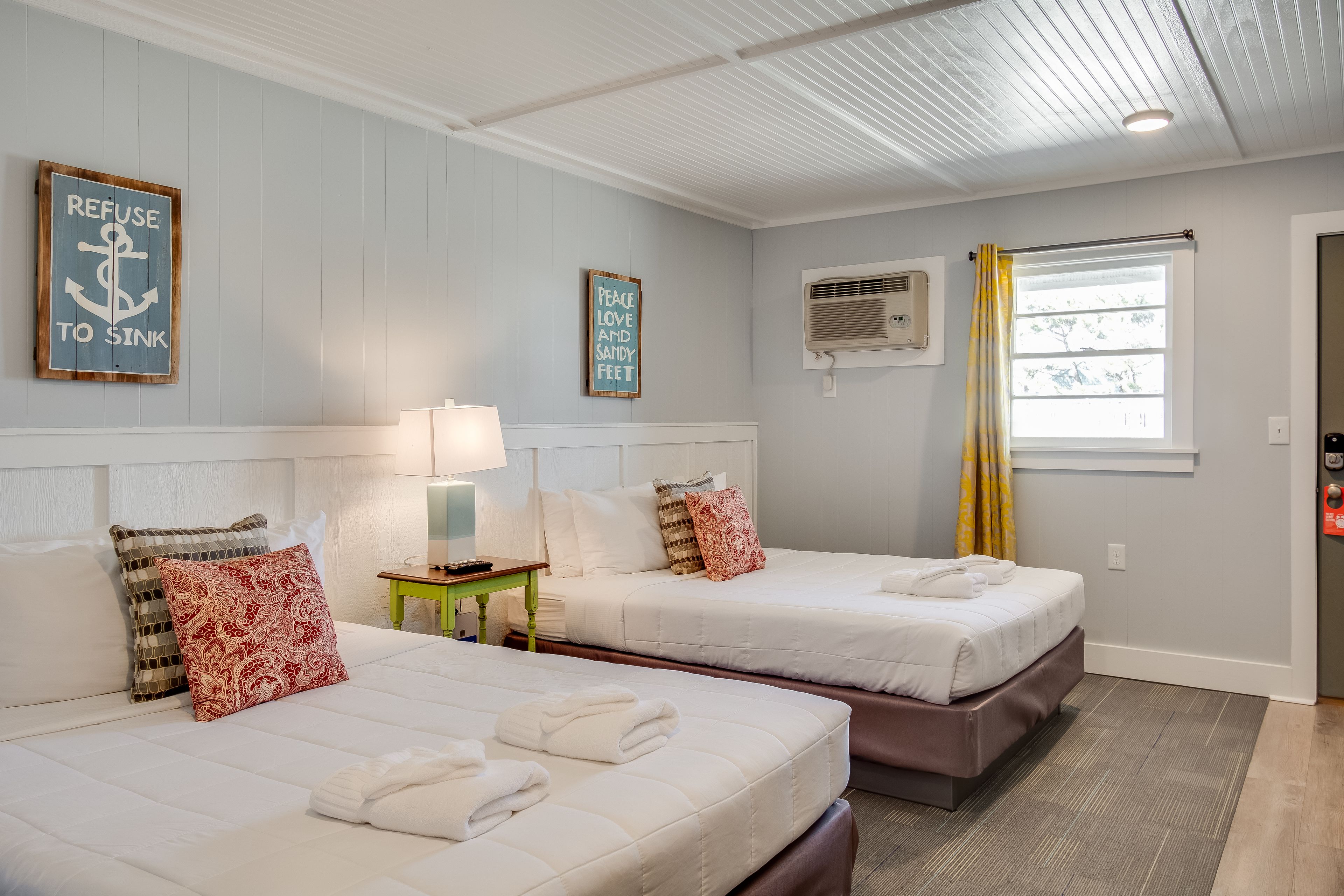 A hotel room with two beds, a small table with a lamp, and a window with curtains.