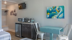 A compact hotel room with a kitchenette, a bed with striped bedding, a teal table, and a colorful abstract painting on the wall. - Tar Heel Motel (Nags Head)
