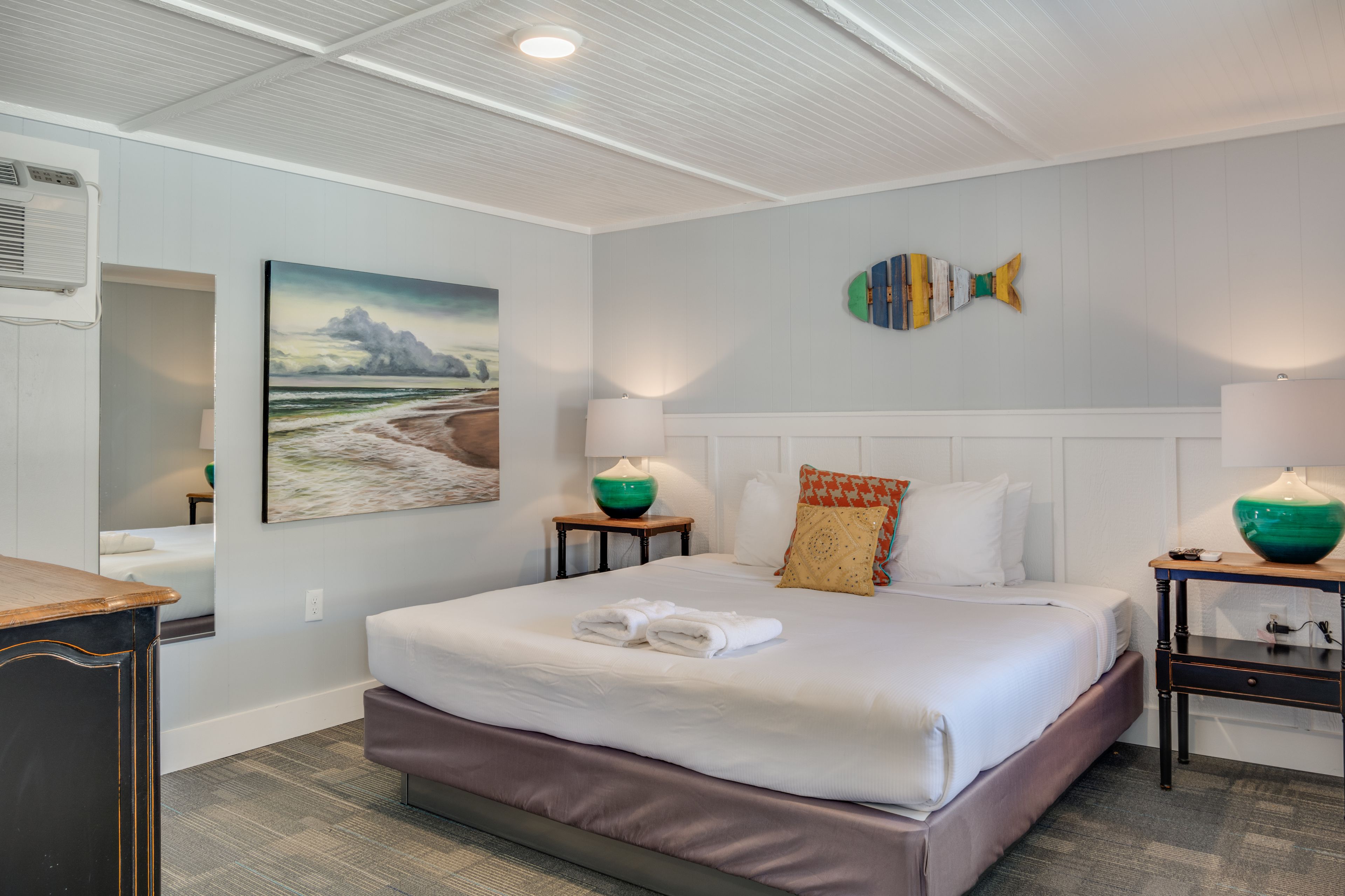 Habitación de hotel con una cama grande, dos mesitas de noche, un adorno de pez montado en la pared y un cuadro de una playa.